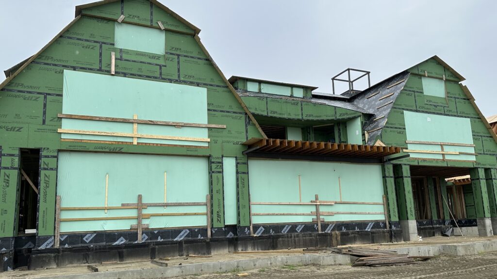 bioaqualife: A partially constructed house with exposed green sheathing, wooden framing, and several openings for future doors and windows. The roof is unfinished. Scaffolding and construction materials are visible around the building site. The ground is a mix of dirt and concrete.