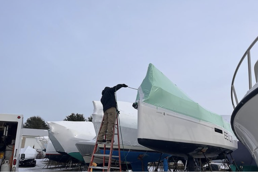 bioaqualife: A person stands on a red ladder while covering a yacht with a green protective cover, made from biowrap. Other boats in the background are covered with white protective covers. Snow-covered ground and trees can be seen in the distance under a cloudy sky, showcasing effective winterization with eco-friendly materials.