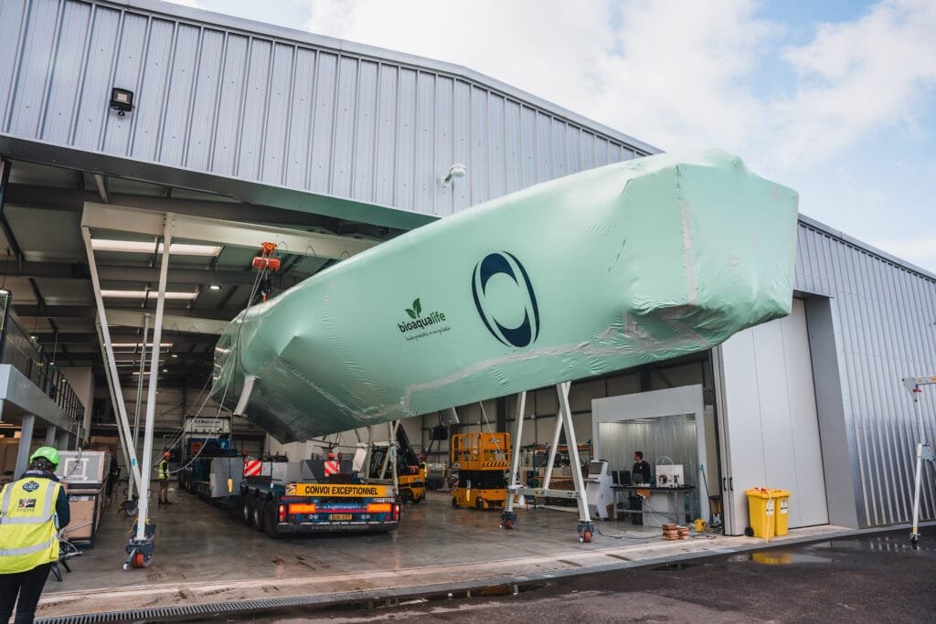 bioaqualife: A large, mint-green boat is being moved out of a modern industrial shed using a blue transport vehicle. Workers in safety gear are visible around the boat. The boat has a logo with a green leaf and a text that reads "bioplasmarine" on its side.