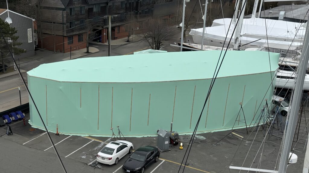 bioaqualife: A large boat covered in a green protective tarp is parked in a lot near some buildings and other boats. Several cars are parked nearby, and there are ladders and equipment around the boat. The tarped boat dominates the image with its light green covering.