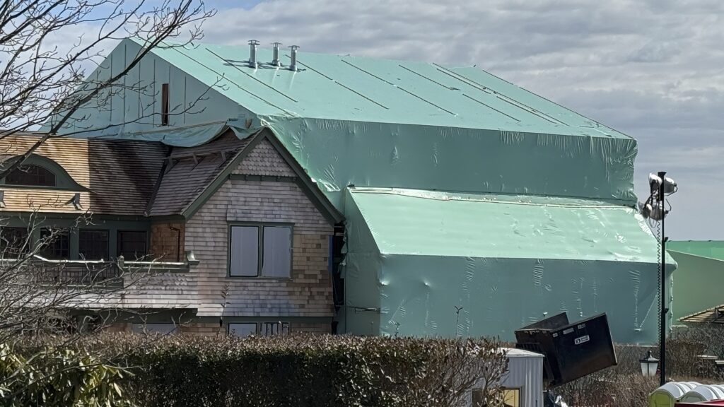 bioaqualife: A large house is partially covered in green construction sheeting, indicating ongoing renovation or construction work. The surrounding area includes some foliage and several pieces of construction equipment. The sky is cloudy.