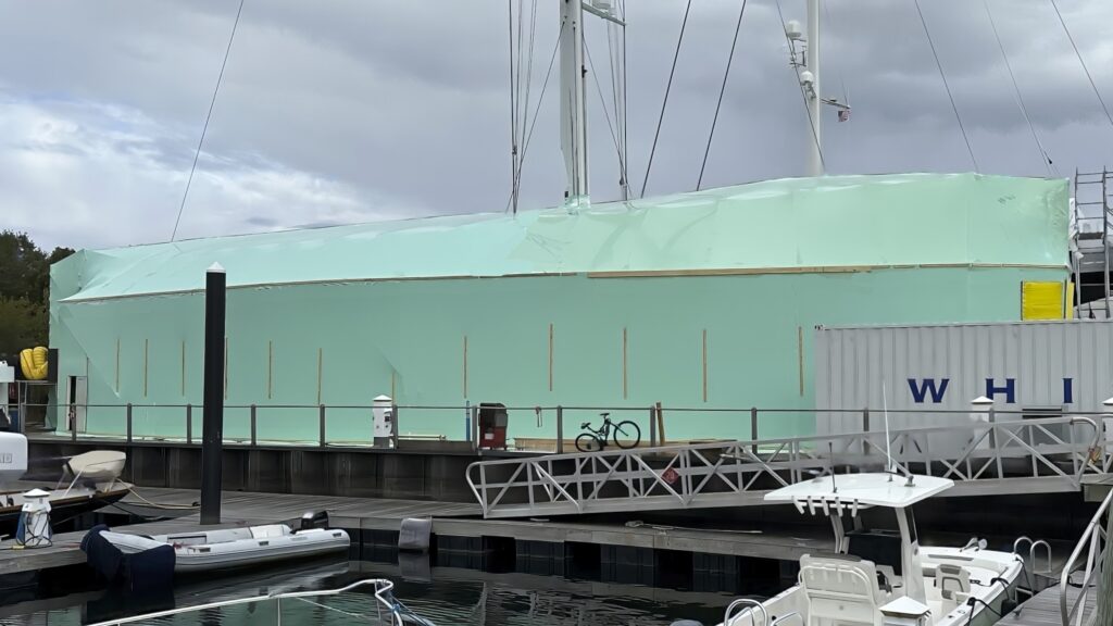 bioaqualife: A large boat covered in green protective sheeting is docked at a marina. A bicycle is parked beside the dock, and several smaller boats and yachts are visible in the foreground. A cloudy sky looms overhead.