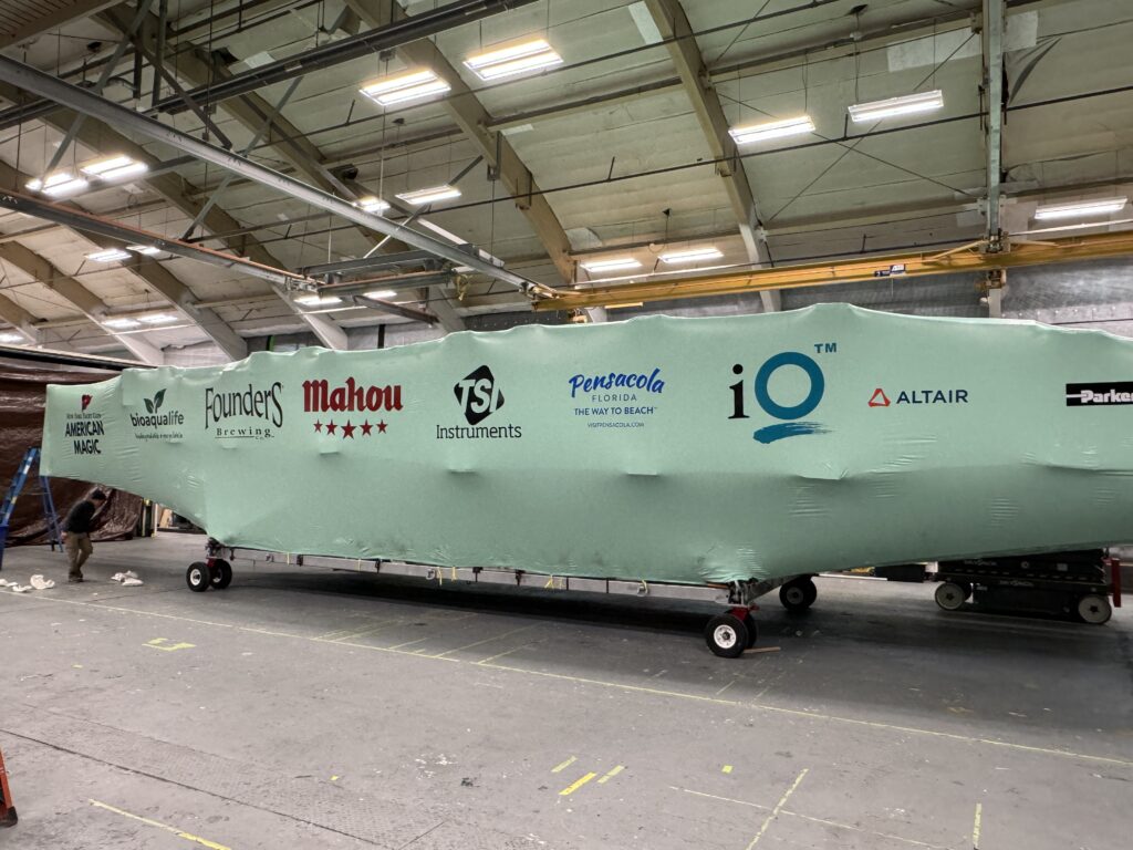 bioaqualife: A large object wrapped in green protective covering inside a spacious warehouse. The covering displays several logos, including American Magic, Founders Brewing, Mahou, Pensacola, IQ Technology, Parker, and others. The object is mounted on a wheeled platform.