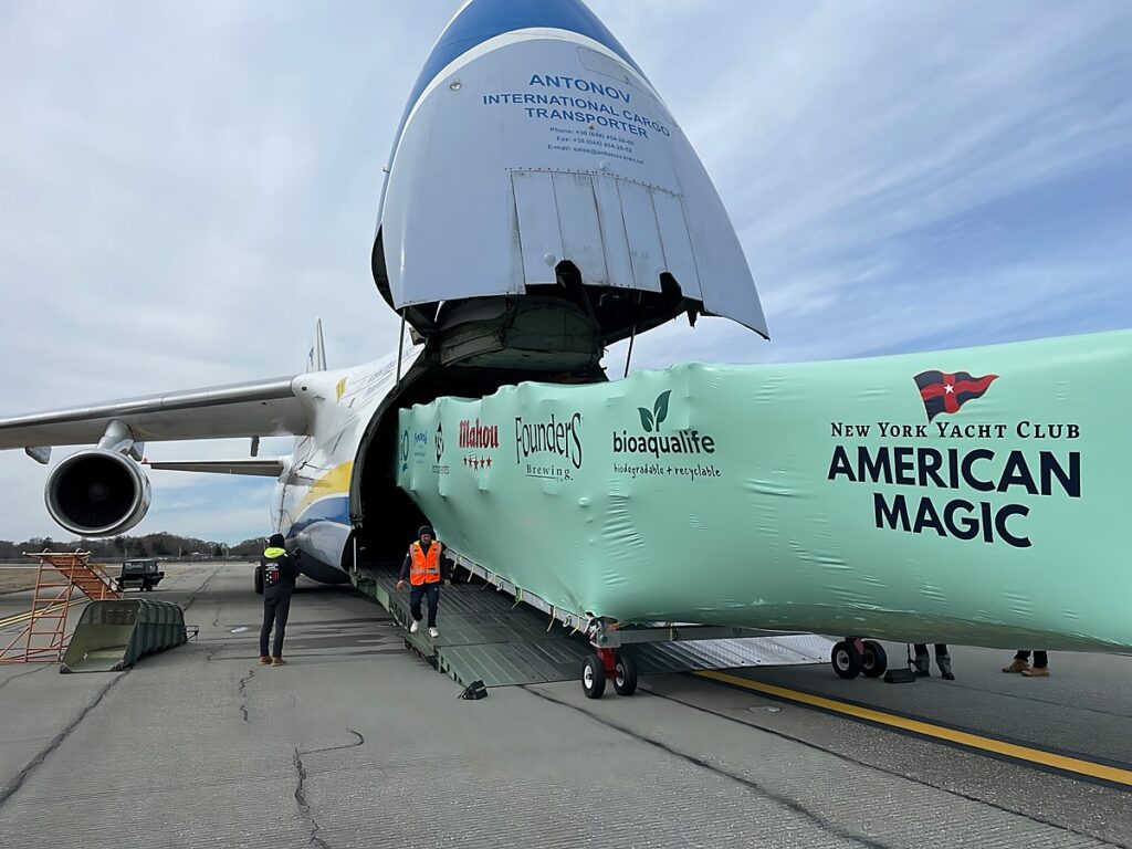 bioaqualife: A large Antonov cargo plane is being loaded with a green-wrapped yacht branded with "New York Yacht Club American Magic" and various sponsor logos. The plane's nose is lifted open, and workers in high-visibility clothing are assisting with the loading process.