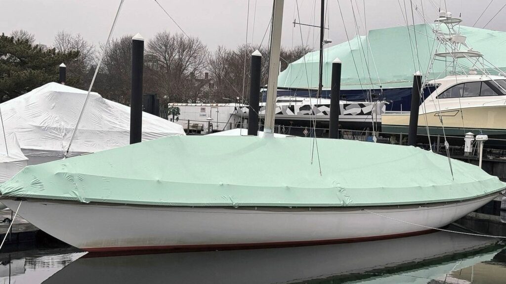 bioaqualife: A sailboat covered in a light turquoise tarp is docked at a marina. Nearby, another boat is also covered, part of it visible on the left side, while more boats can be seen in the background under grey, overcast skies.