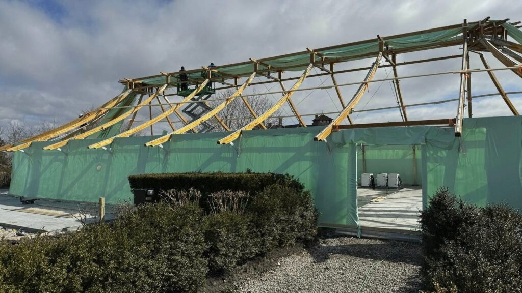 bioaqualife: An unfinished building structure with visible wooden beams and green construction mesh. The roof is incomplete, and there's construction equipment inside. Bushes are seen in the foreground, while the background shows a cloudy sky.