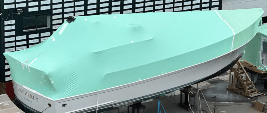 bioaqualife: A large boat is covered with a green protective tarp. It's positioned in a shipyard or storage area, elevated on stands. Nearby, another covered boat and industrial building are partially visible.