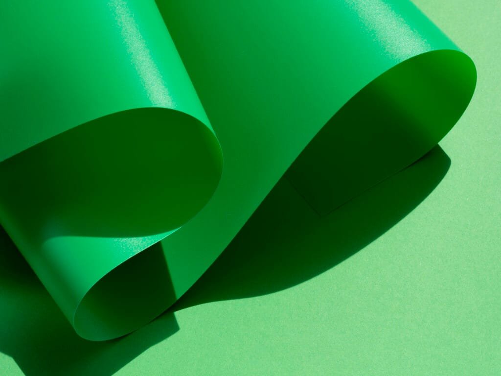 bioaqualife: A smooth, glossy green sheet of paper rolled in a wave pattern, casting shadows on a lighter green surface. The light highlights the texture and shape of the paper, creating a dynamic contrast with the background.