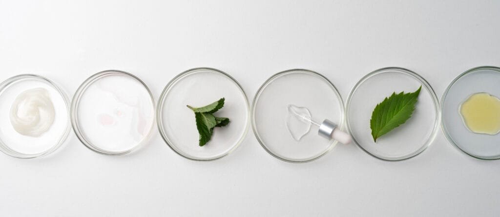 bioaqualife: A row of six circular glass dishes on a white surface containing different skincare ingredients: cream, liquid, two green leaves, a clear serum with a dropper, and another liquid.