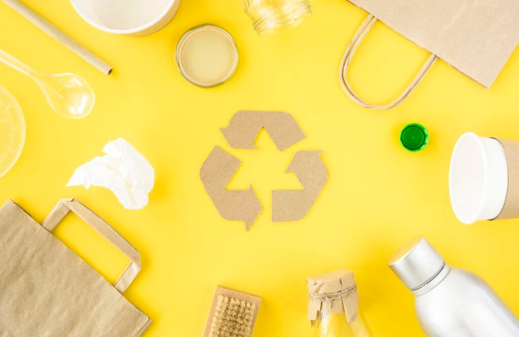 bioaqualife: A recycling symbol made of cardboard is surrounded by various eco-friendly items, including a reusable bottle, shopping bag, glass jars, and biodegradable utensils on a yellow background.