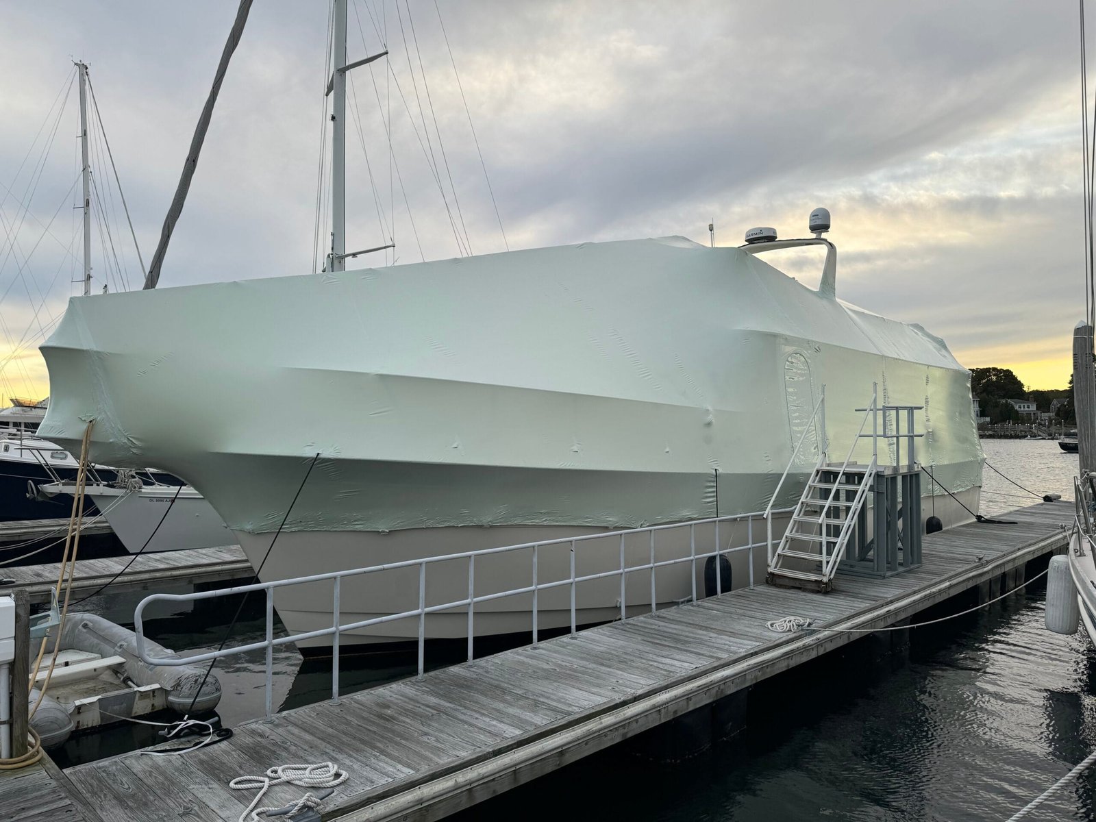 bioaqualife: A large yacht completely covered with a protective wrap is docked at a marina. The dock has narrow paths and ladders, with a cloudy sky in the background. Other boats are faintly visible nearby.