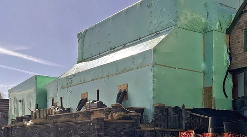 bioaqualife: A large building covered in green protective tarpaulin, with several black vent pipes protruding. The base is surrounded by stone walls and some construction materials. The sky is clear and blue.