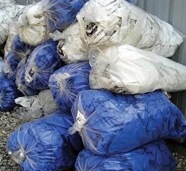 bioaqualife: A stack of large plastic bags filled with blue and white fabric materials. The bags are tightly sealed and piled in an outdoor setting. The ground is covered with small rocks, and a metal structure is visible in the background.