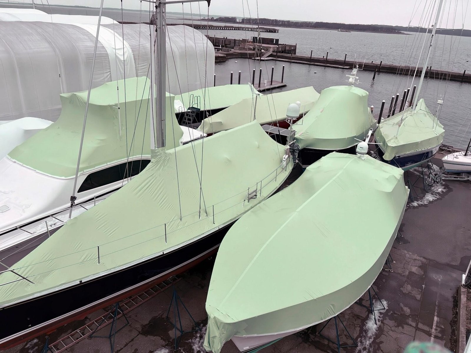 bioaqualife: Boats covered with green tarps are docked at a marina. The marina, surrounded by a calm body of water, has several docks visible in the background. The scene suggests these boats are being stored or protected for the off-season.