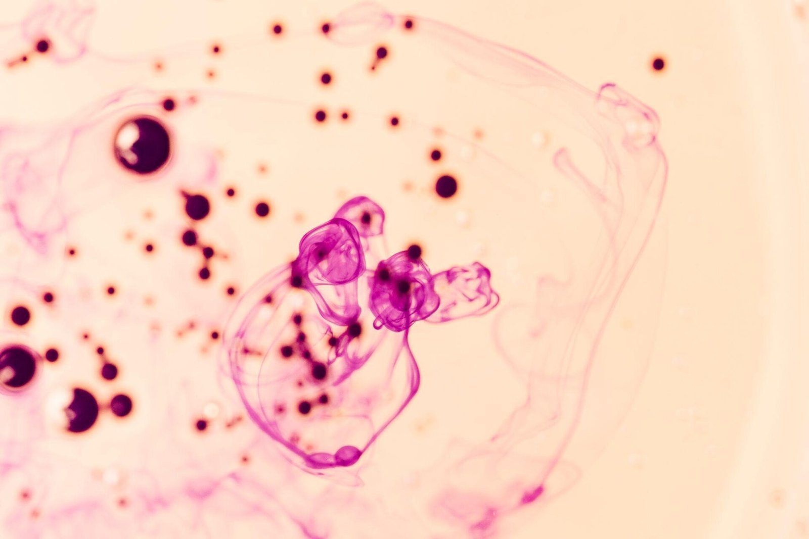 bioaqualife: Abstract image of pink ink swirling in liquid, with dark pink and black ink droplets scattered on a light beige background. The ink creates flowing, cloud-like patterns and circular shapes.