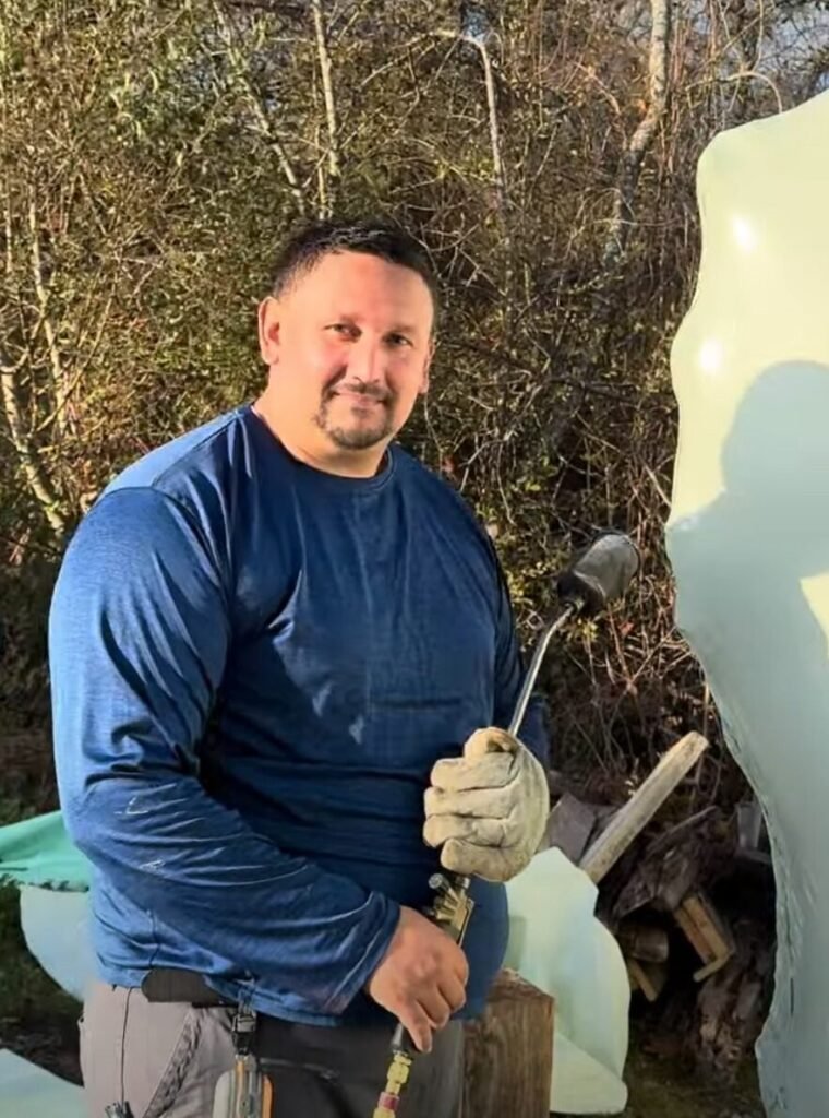 bioaqualife: A man in a blue long-sleeve shirt and work gloves holds a blowtorch outdoors, standing next to a large block of ice with trees and branches in the background.
