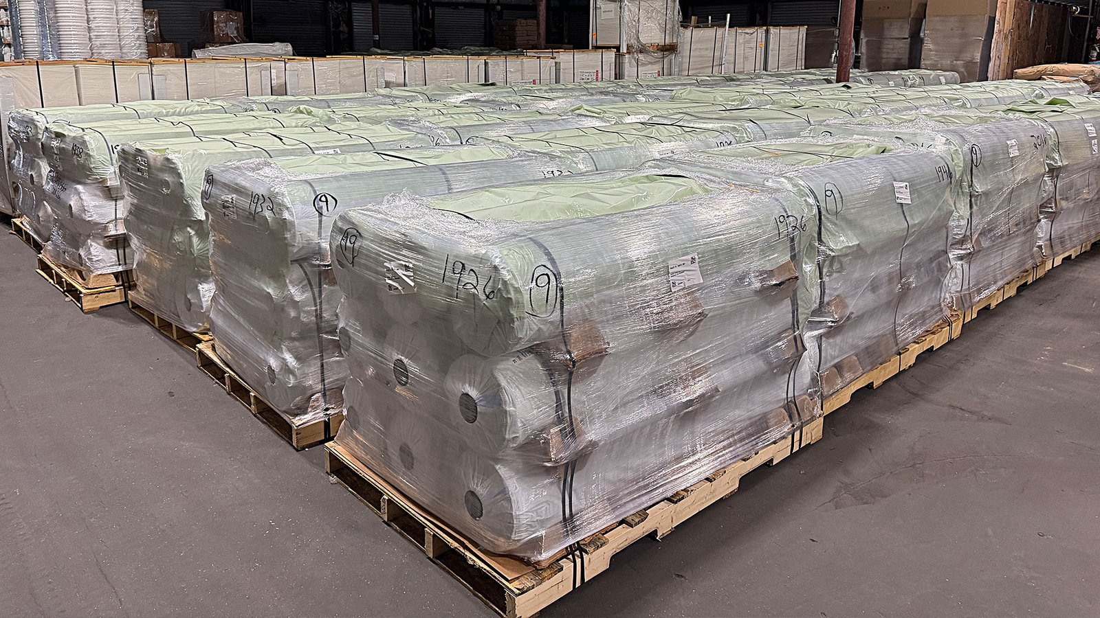 bioaqualife: Stacks of large, tightly wrapped rolls of material on wooden pallets are arranged in rows inside a warehouse with a concrete floor and industrial shelving in the background.