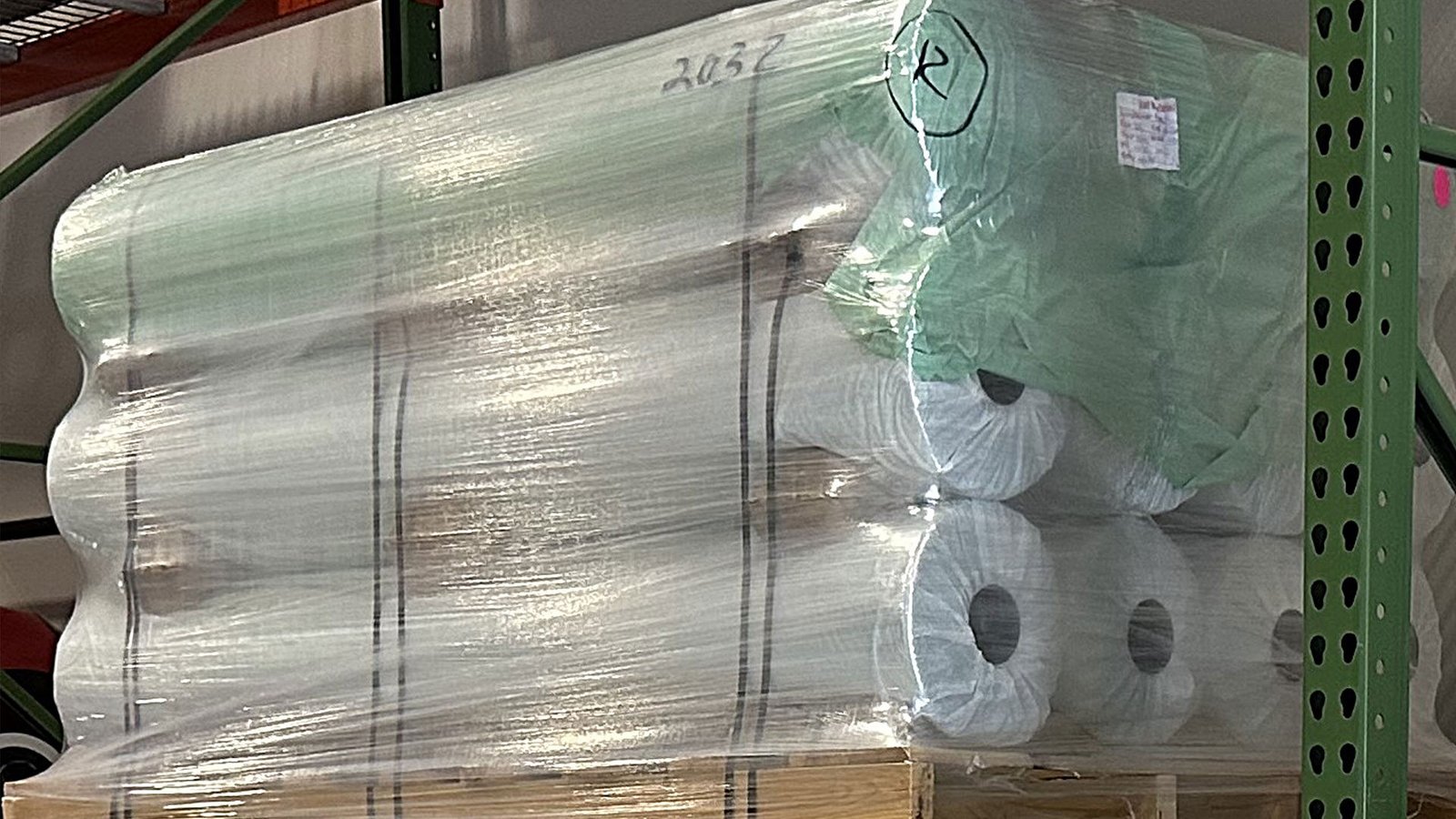 bioaqualife: A stack of large rolls of white fabric wrapped in clear plastic sits on a wooden pallet in a warehouse storage rack. Handwritten markings and labels are visible on the packaging.